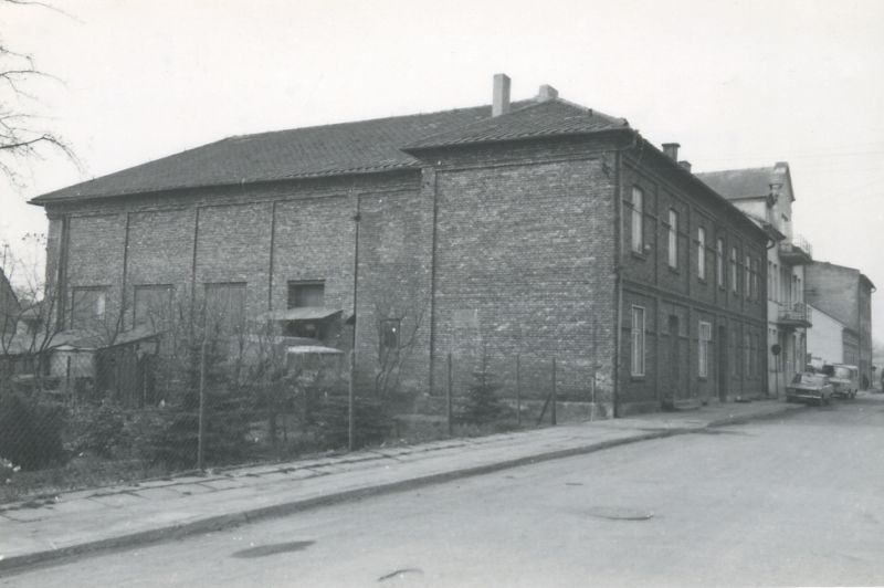 Large brick building by an asphalt street. Two cars parked on the street.