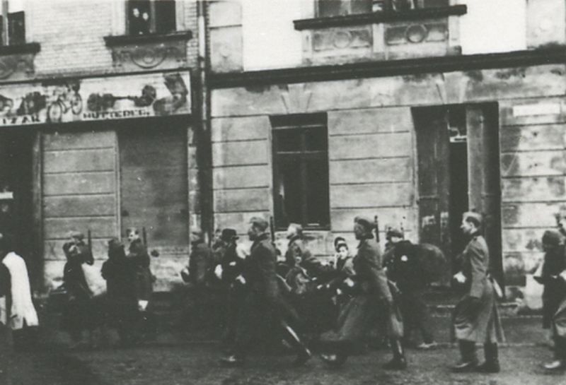 Displacements from Oświęcim. Men in coats, women and children with luggage and bundles are walking down the street, guarded by German policemen in uniforms.