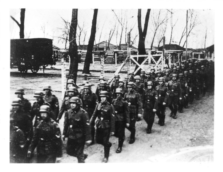 SS men from KL Auschwitz crew wearing uniforms and helmets on their heads, marching next to a warehouse of construction materials. In the distance, a freight wagon standing on a siding and buildings.