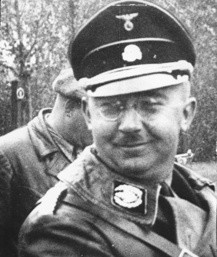 Heinrich Himmler.
A middle-aged man in a German SS uniform, wearing a cap with a skull and glasses, sitting in a car, smiling.