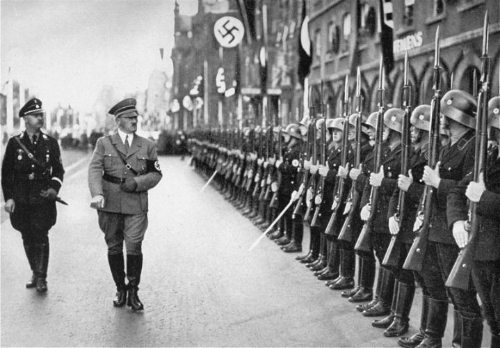 Two middle-aged men in German uniforms: Heinrich Himmler and Adolf Hitler. In front of them, German soldiers in uniforms and helmets presenting their weapons. A flag with a swastika hanging on the tenement house behind them.