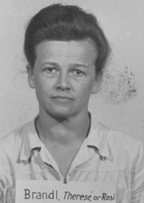 Process photo. Woman in a blouse with collar and pinned back hair. She has a plaque with surname around her neck.