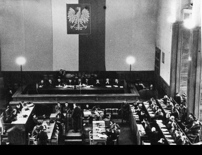 The courtroom consists of prosecutors, defendants and defense attorneys.  Above them, a Polish flag and an emblem hanging vertically on the wall.