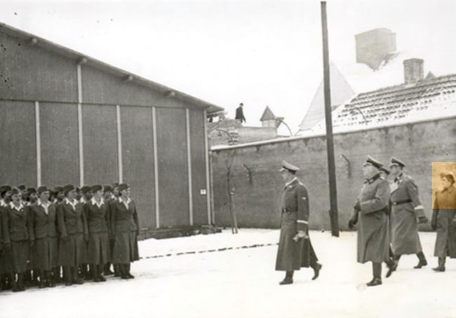 On the left side, supervisors in service uniforms standing in rows.  On the right, three SS men and a supervisor approaching them. Winter. In the background, someone working on the roof of the building.