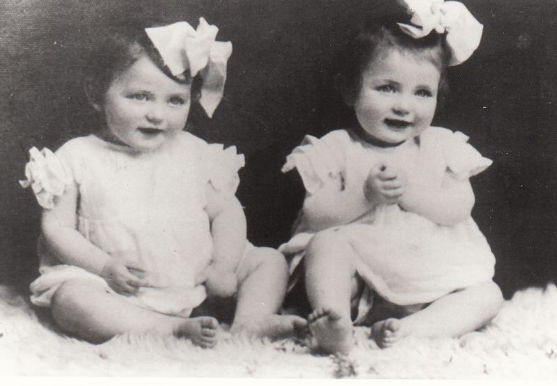 Sitting, smiling girls, around one year old, wearing dresses, with bows in their hair.