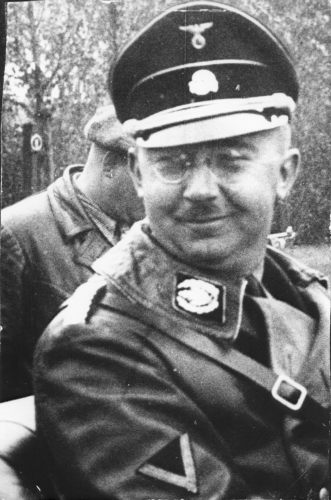 Portrait of a man in round glasses, a military uniform and a cap.  On the Totenkopf cap: skull and crossbones. Smiling, relaxed. In the background a silhouette of another person, a forest.