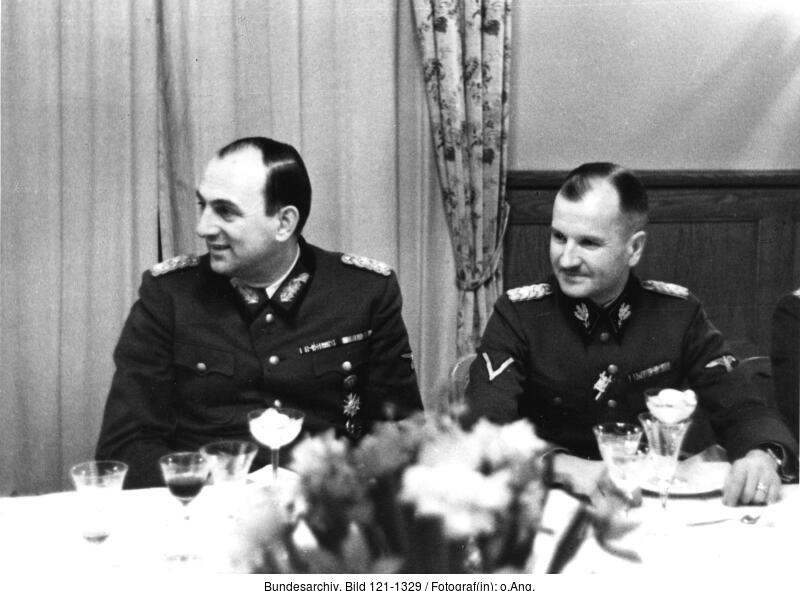 Two middle-aged men sitting at a table. They are talking and smiling. They are dressed in uniforms. Plates and numerous glasses on the table. In the background there is a wall, curtains and wood paneling.