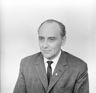 Portrait of a middle-aged man.  Dressed in a suit, a tie and a shirt. Gazing to the left side. There is a pin in the lapel of the jacket.