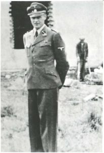 A photo of the silhouette of a middle-aged man in a German uniform. He is looking from the side towards the camera lens. In the background there is a building and a person standing nearby with crutches.