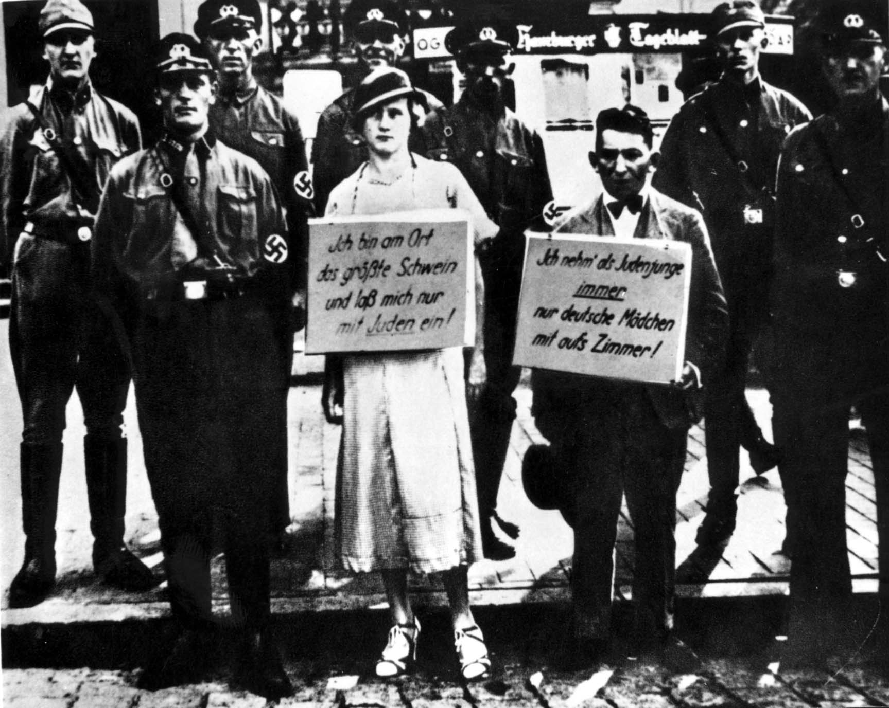 A man and a woman standing in the street, with signs hanging from their chests. They are elegantly dressed, the man in a suit, the woman in a dress and wearing a hat. Behind them are men in uniforms with swastikas on their shoulders.