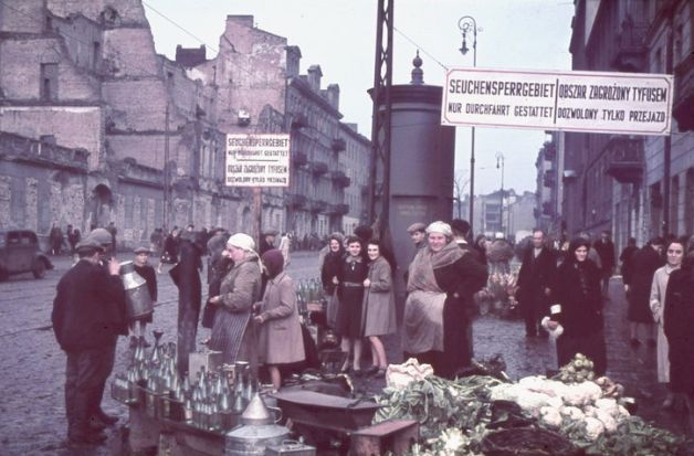 Street stall on a street of Warsaw. Bottles and vegetables displayed at the stall. Older women in aprons and headscarves selling goods. On the street there are men, women and children in coats and hats, some have armbands (with the Star of David) on their coat sleeves.