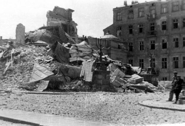 The ruins of the Great Synagogue in Warsaw with a huge Jewish candlestick in the middle, rubble behind it, a tall building on the right side of the square. Two boys are leaning against the railing opposite the ruins.