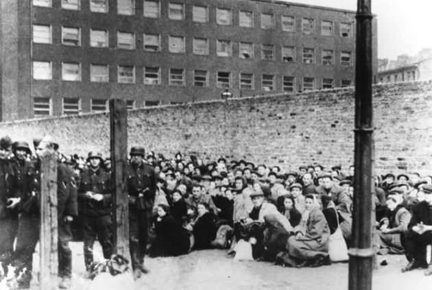 A crowd of people sitting in the square, men, women and children in coats and with bundles. They are guarded by German soldiers in uniforms. A wall behind them.