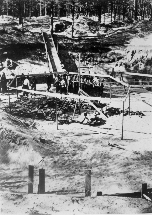 Grupa ludzi prowadzona przez żołnierzy niemieckich z karabinami na miejsce egzekucji. Wokół doły wykopane w ziemi i las.