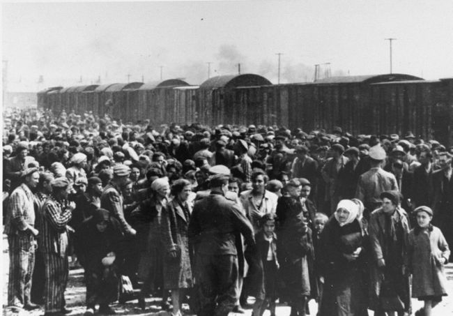Tłum ludzi: mężczyźni, kobiety, dzieci, ludzie starsi stojący na rampie kolejowej z Birkenau. Na pierwszym planie esesmani w mundurach oraz kilku więźniów w pasiakach. Na rampie stoi pociąg towarowy. W oddali fragment bramy wjazdowej do Birkenau. 
