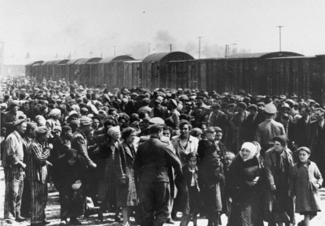 Tłum ludzi: mężczyźni, kobiety, dzieci, ludzie starsi stojący na rampie kolejowej z Birkenau. Na pierwszym planie esesmani w mundurach oraz kilku więźniów w pasiakach. Na rampie stoi pociąg towarowy, w oddali widoczny fragment bramy wjazdowej do Birkenau.