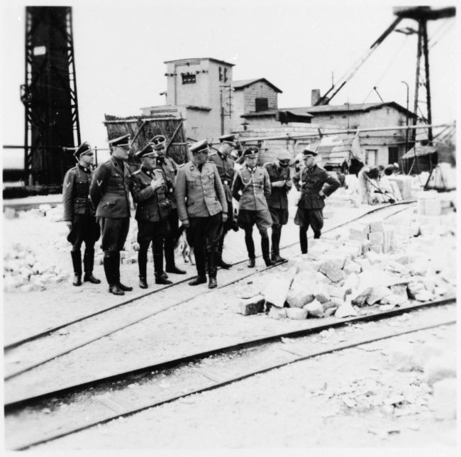 Esesmani w mundurach wizytujący kamieniołom. Na pierwszym planie tory kolejowe i bloki kamienne, w tle zabudowania.