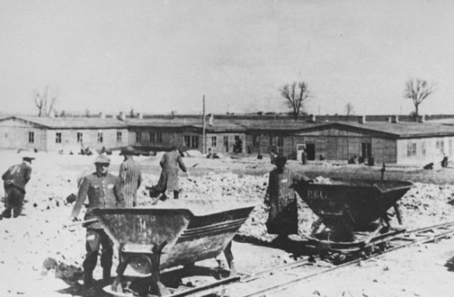 Więźniowie w pasiakach przy pracy przy załadunku kamienia. W tle drewniane baraki obozowe. 