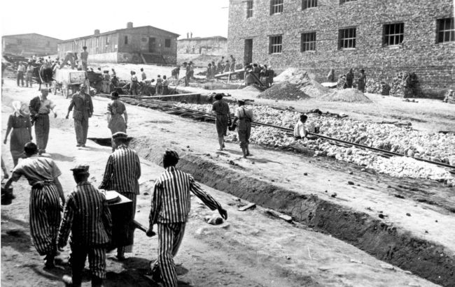 Więźniowie i więźniarki przy pracy przy torach kolejowych. W tle murowany budynek z kratami w oknach oraz drewniane baraki obozowe. 