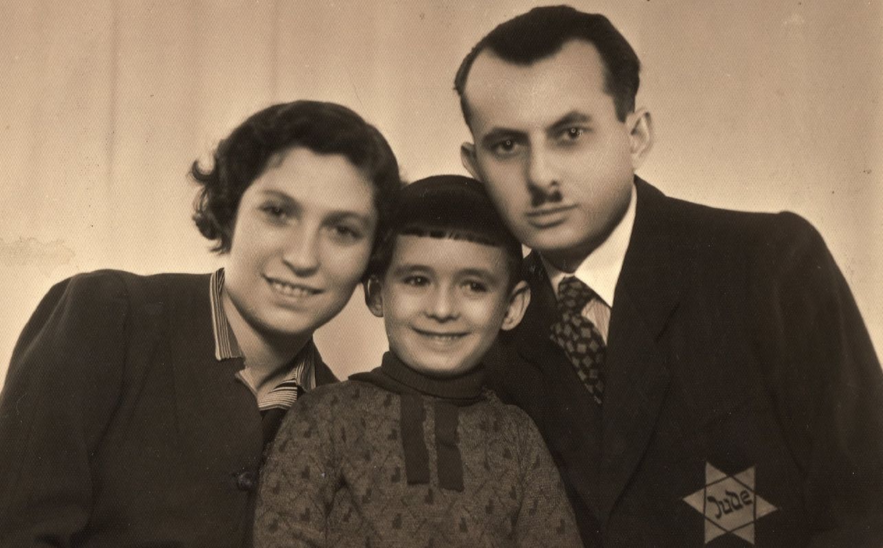 Portrait of a married couple with a baby boy. Their heads are turned towards each other. Faces smiling, gaze directed towards the lens. The family dressed elegantly, the man in a suit with a star of David sewn on it with the word 