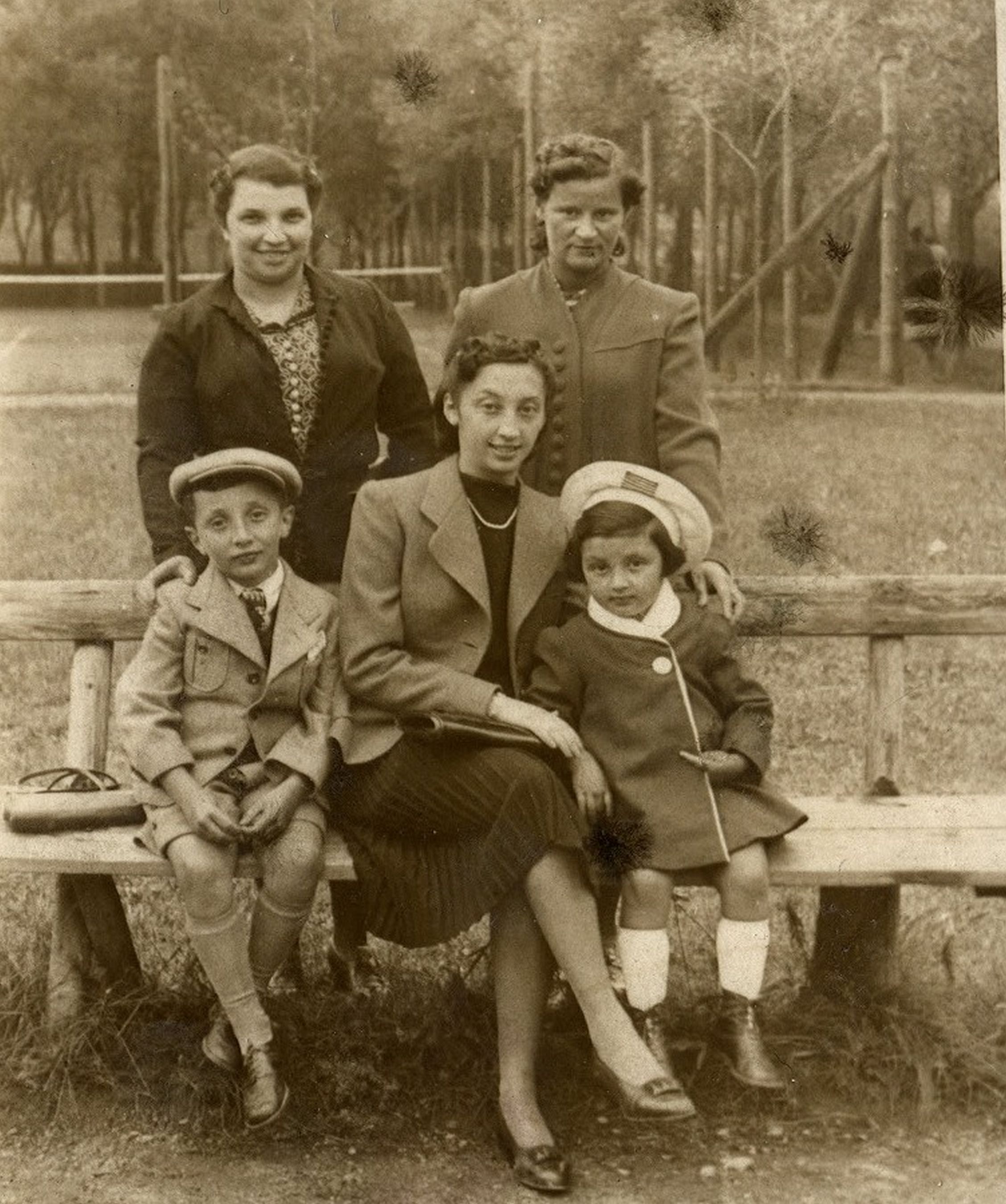 A woman and two children sitting on a bench. Behind them two women. In the background a meadow, a fence, trees.