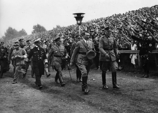 In the foreground Adolf Hitler walks together with his comrades. In the background, crowds gathered on the hill - they perform the Nazi salute (Heil Hitler).