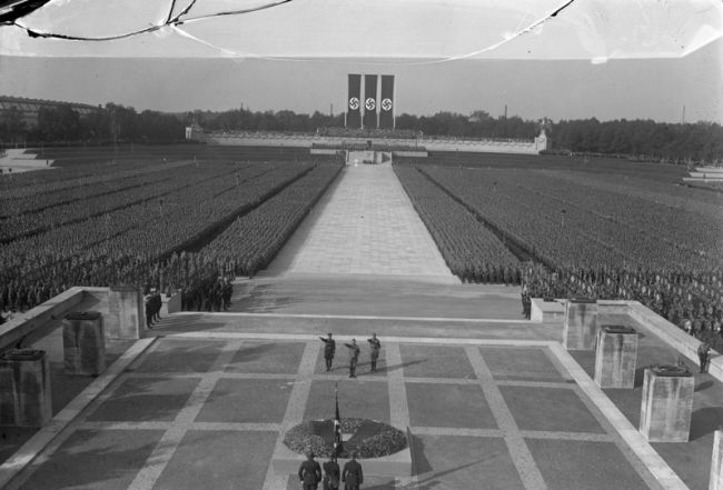 To the right and left of the photo are tens of thousands of assembled NSDAP party members, standing in even rows. In the middle, an empty strip of wide sidewalk. In the background are three oblong hanging Nazi flags. In the foreground at the bottom three NSDAP members.