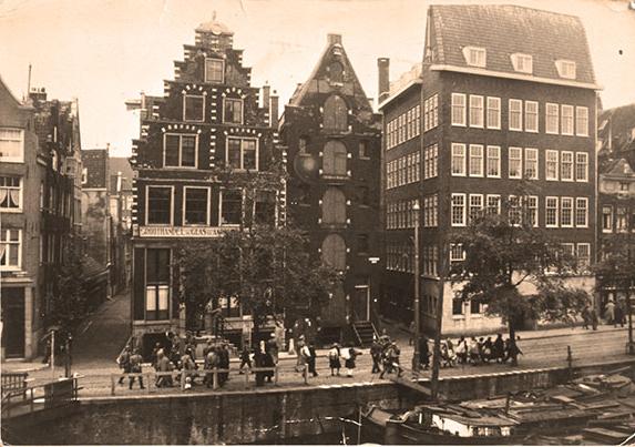 A street in the Netherlands. A group of people standing on a high, concrete bank, being herded towards the SS men. In the background there are several stories high tenement houses. There is a river flowing below, moored barges.