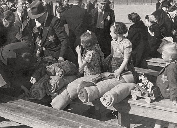 A group of people – men, women and small children –sitting on wooden benches. Next to them, rolled up blankets and a wooden toy - a horse on wheels. A crowd of people in the background.