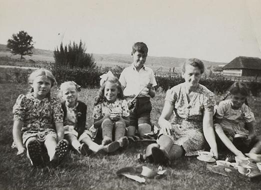 A woman and five children sitting on the grass.  The horizon behind them, a wooden thatched cottage on the right, in the background.