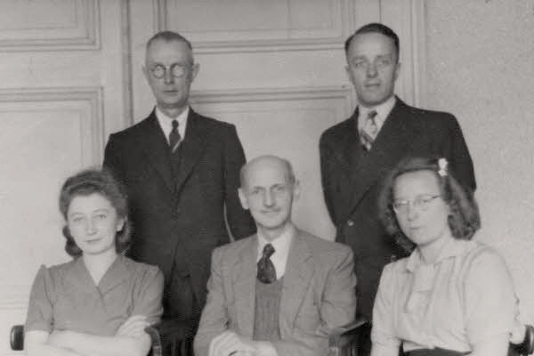 A group of people posing for a photo indoors. Two women are sitting and there is a man in the middle. Two men are standing above them. All middle-aged. Faces focused, serious. A tall door in the background.