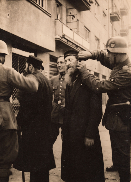 Na pierwszym planie żołnierz niemiecki obcina brodę żydowskiemu mężczyźnie. Obok żołnierz niemiecki patrzy i się uśmiecha. Po lewej stronie odwrócony plecami mężczyzna, prawdopodobnie Żyd, rozmawia z żołnierzami niemieckimi.  