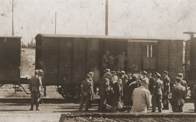 Stacja kolejowa. Przed wagonem towarowym stoi tłum ludzi. Pojedyncze osoby wsiadają do wagonu. Pilnowany jest przez policję porządkową oraz żołnierzy niemieckich. 