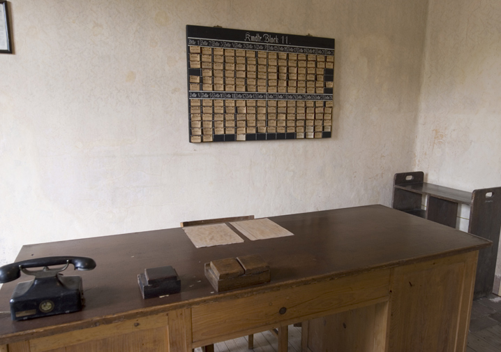 Room of the manager of block 11 (Blockführer). On the wall a blackboard with listed cells located in the basement of the block and sheets of paper with the names of the prisoners in each of them. The room still has a desk, telephone, documents and a chair.