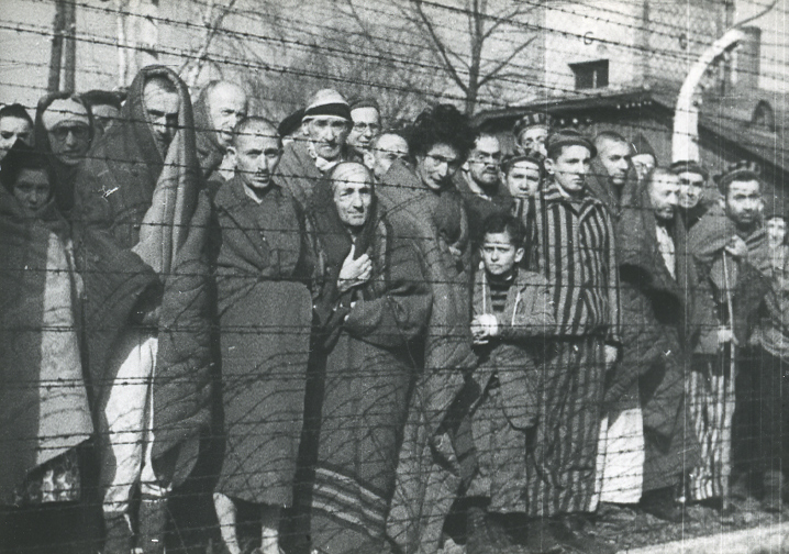 Auschwitz I. A group of liberated prisoners pose from behind the wires of the camp fence. Dressed in striped uniforms - prisoners clothing, some people are covered with blankets.