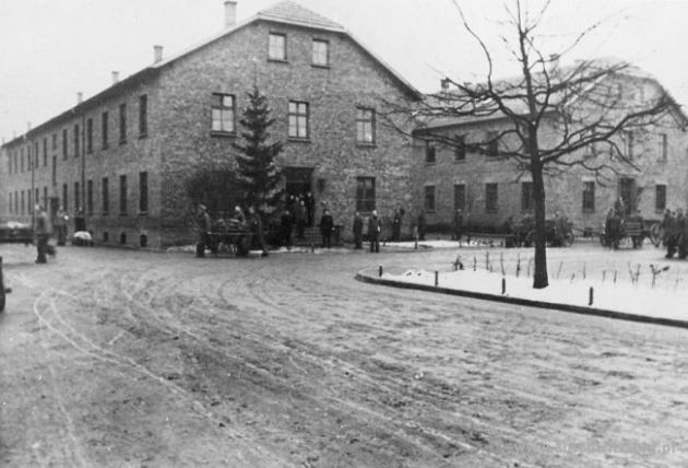 Front of a single-storey brick-walled building with sloped roof. A tree in front of the building, prisoners in striped uniforms are walking by.