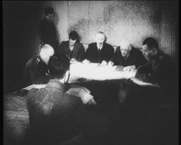 Men sitting at a table bent over documents in focus. In front of them a man with his back turned to the photographer.