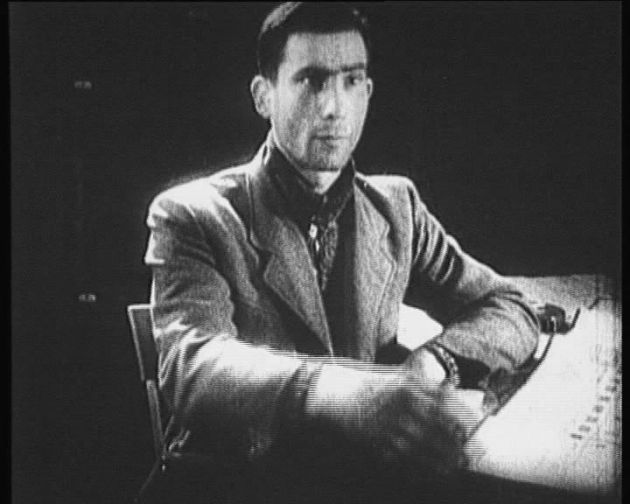 A man is sitting at a table. Face focused, serious expression. Dark hair. A jacket and a shirt.