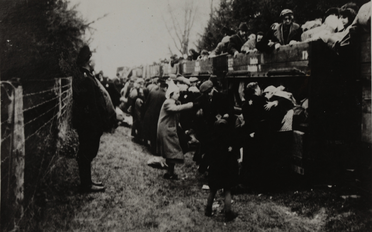 On the right, coal cars filled with people going to the extermination camp. Some people are still boarding. An obese soldier in a uniform and a cap is watching the situation.