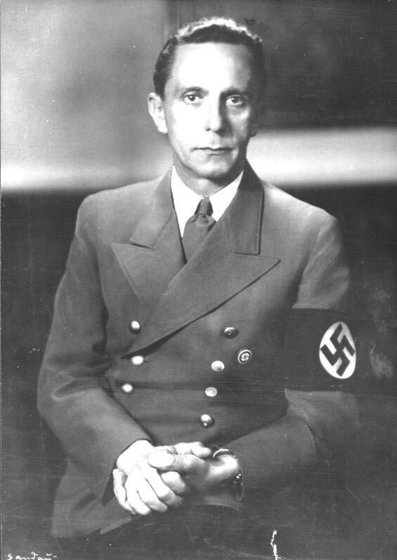 Portrait photograph of a man wearing a double-breasted suit with gold buttons. On the sleeve of the left arm an armband with a swastika. Face focused, directed towards the camera. Serious expression, lips pursed.