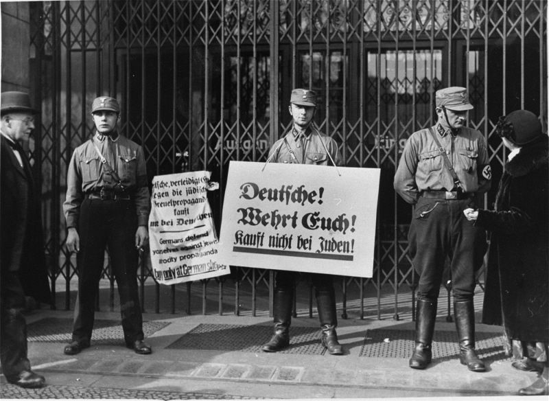 Three men dressed in SA uniforms standing in front of a shop window closed behind bars. One of them is holding a banner that reads: 