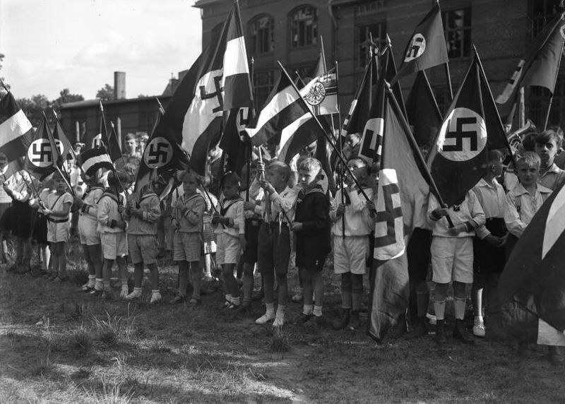 A group of children - several-year-old each - dressed in navy clothes. They are holding small bannerets with Nazi flags - swastikas.
