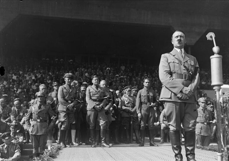 In the foreground, on the right Adolf Hitler standing and speaking in front of a microphone. In the background officers and soldiers on the stands behind him.
