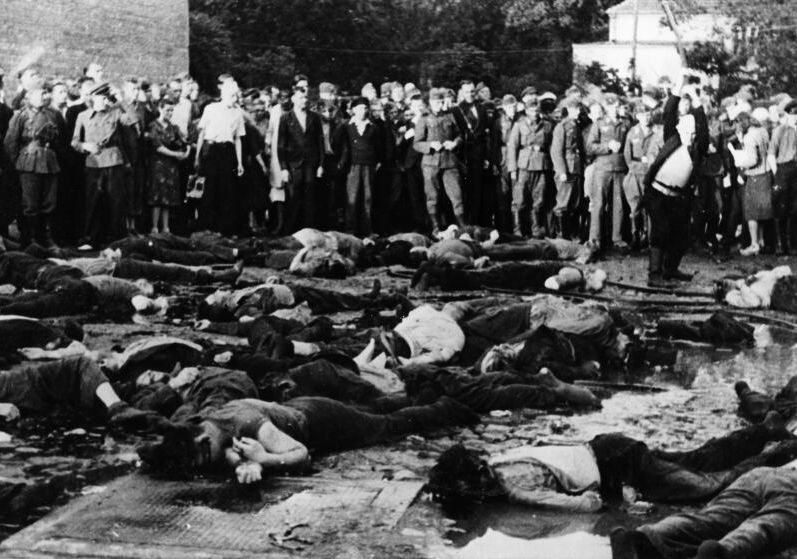 A moment just after the massacre. In the foreground bodies of the victims. In the background above them a crowd standing and watching. On the right a man preparing to hit another man lying down, presumably wanting to hit and kill him.