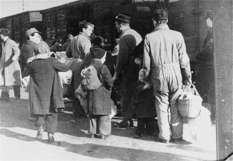 Station platform, adults and children with their backs turned are about to board freight trains.