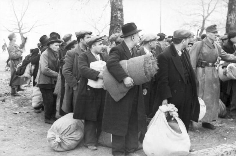 A group of men in coats and winter clothes standing and waiting to be directed to the train. They are holding luggage in their hands or on the ground in front of them. They are being led by soldiers.