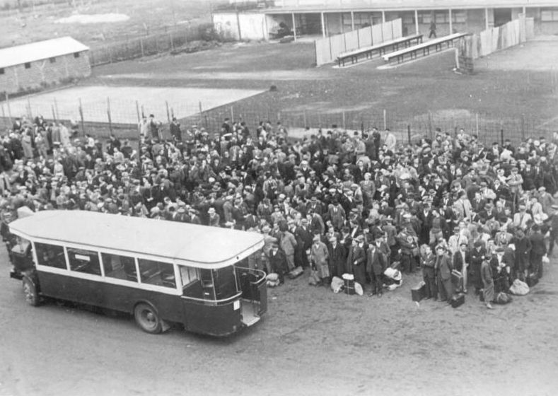 A crowd of people standing and waiting in front of the bus. In the background most likely a transit camp - a wire fence, benches, buildings.