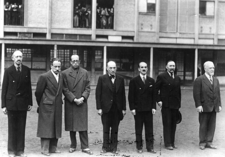 Seven middle-aged men dressed in suits, hats and coats are standing in front of a building. In the windows of the vast building stand people that are watching them.