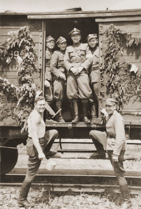Polish soldiers standing in the doorway of a freight carriage which is decorated with shrubs and flowers.