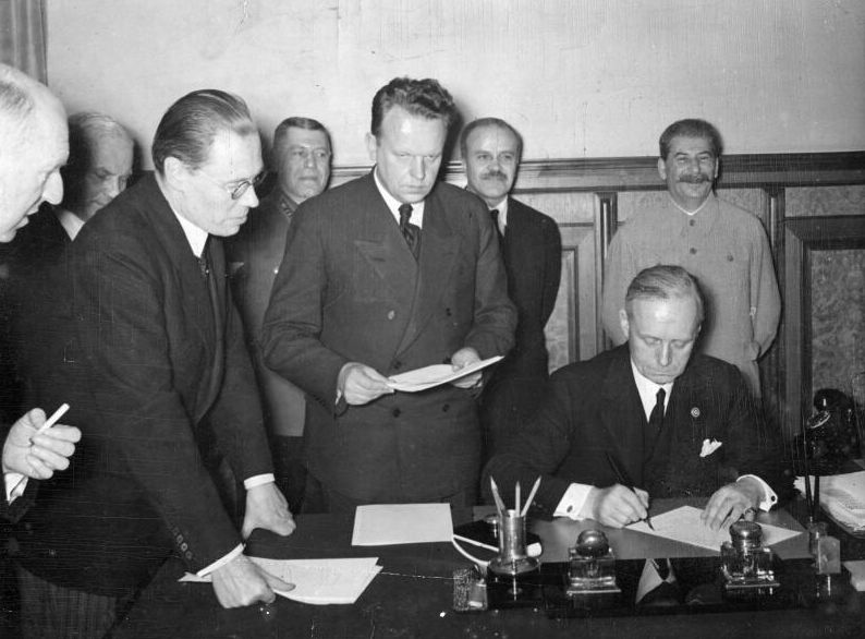 A man sitting at a desk signing a document. Just beside him are two men standing over the desk. One is holding the document. Behind them, the other men are watching - among them smiling Joseph Stalin.